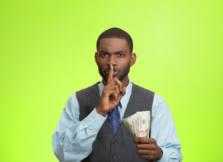 Closeup portrait handsome corrupt guy businessman holding dollar bill in hand showing shhh sign finger to lips isolated green background. Bribery concept politics, business diplomacy. Face expressionの写真素材