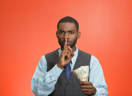 Closeup portrait handsome corrupt guy businessman holding dollar bill in hand showing shhh sign finger to lips isolated red background. Bribery concept politics, business diplomacy. Face expressionの写真素材