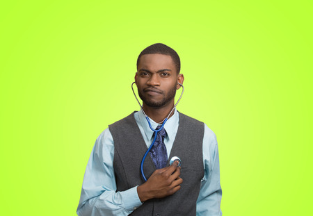 Closeup portrait serious executive man, business person, worker listening to his heart with stethoscope isolated green background. Preventive medicine, financial condition concept. Face expressionsの写真素材