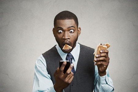Closeup portrait surprised funny looking corporate business man holding, reading bad news on smart phone while eating cookie, about to choke isolated grey wall background. Face expression, emotionの写真素材