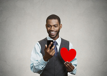 Closeup portrait cheerful young man checking his smart phone, holding red heart in his hand, isolated grey wall background. Human facial expressions, emotions, feelings, body language, life perceptionの写真素材