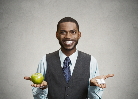 Closeup portrait young fit happy executive man holding green fresh apple in one hand, pills, vitamins in another, trying to decide which choice is the best one. Face expressions, emotions, health careの写真素材