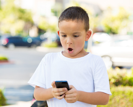 Portrait child, surprised boy, little man texting on mobile using smart phone isolated outdoor, outside background. Funny looking human face expression, emotion, reaction, body language. Communicationの写真素材