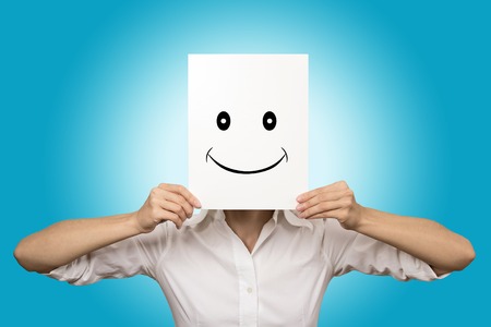 Woman showing happy emotion holding covering front of face with smiling paper mask isolated on blue background. Human facial expressions, fake identity, unknown, behind the scene conceptの写真素材