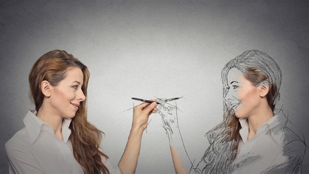 Create yourself, your future destiny, image, career concept. Attractive young woman drawing a picture, sketch of herself on grey wall background. Human face expressions, determination, creativity