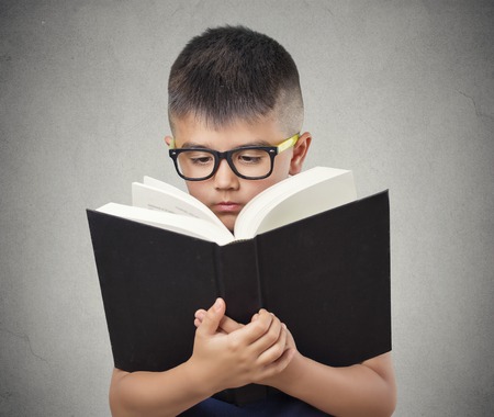 closeup portrait child boy with glasses reading holding book too close having difficulty to see text, isolated grey wall background. Face expressions. Education concept. Vision problems in childrenの写真素材