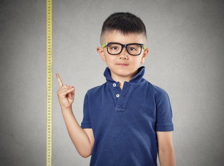 I'm growing up. Child with glasses pointing at his height on measuring tape beside him, grey wall background. Children development concept. Face expressionの写真素材