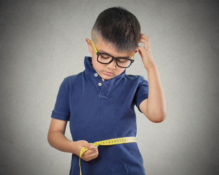 Portrait little boy, child measuring his waistline, scratching head, thinking, isolated grey wall background. Dieting, fitness, healthy nutrition education, body care, weight control conceptの写真素材