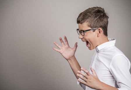 Closeup side view Portrait Angry teenager boy screaming arms up in air isolated grey wall background. Negative face Expression emotion reaction perception body language Conflict confrontation conceptの写真素材