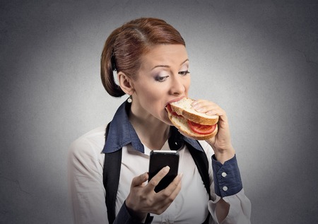 Closeup portrait young serious corporate business woman deal maker reading news message on smart mobile phone holding eating sandwich isolated grey background.の写真素材