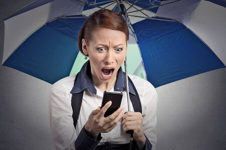 Closeup portrait shocked surprised business woman corporate executive reading bad breaking news on smart phone holding umbrella protected from rain isolated grey background.の写真素材