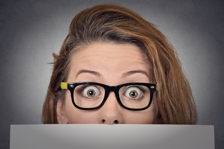 Banner sign woman peeking over edge of blank empty paper billboard. Beautiful young woman with glasses looking surprised and scared funny with wide open eyes isolated on grey wall backgroundの写真素材