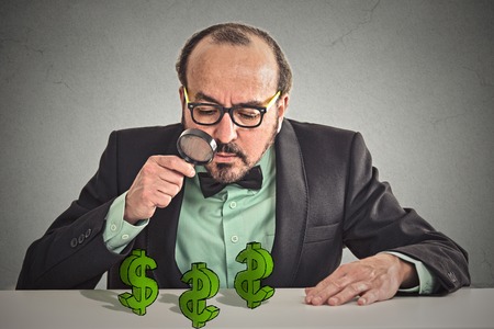 Business man looking through magnifying glass at dollar signs symbol on table isolated grey wall office background.の写真素材