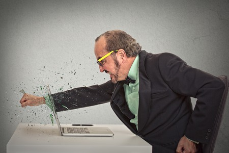 furious businessman throws a punch into computer screaming isolated grey office wall background.の写真素材