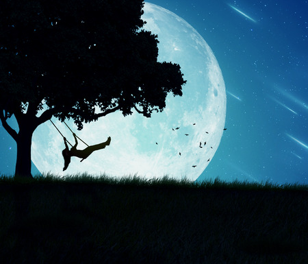 Silhouette of happy young woman on a swing of a tree isolated on beautiful background of moon, earth, night skyline, falling stars. Body vitality, human spirit wellbeing, freedom, happiness conceptの写真素材