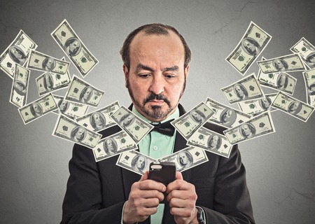 Technology, online banking, money transfer, e-commerce concept. Middle aged businessman in suit and bow tie using smartphone dollar bills flying from screen isolated grey wall office background.の写真素材