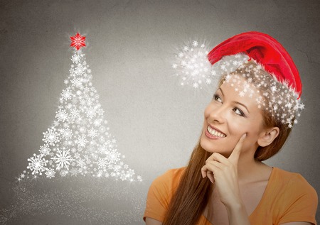 x-mas, winter, happiness concept smiling woman in red santa helper hat looking at christmas tree on grey wall background. Positive emotion face expression perception visionの写真素材