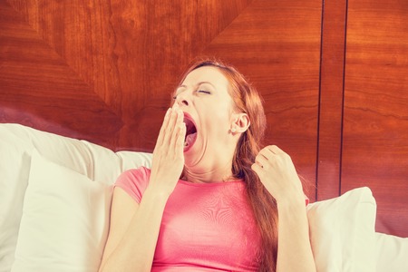Woman yawning and stretching her arms as she lies in bed trying to wake up for a new day. Human face expression emotion body language. Lack of adequate sleep conceptの写真素材