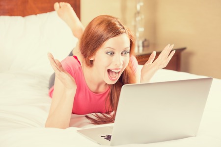 Excited young beautiful woman with arms raised using looking at her laptop screen laying in bed. Positive human emotion happy facial expression, body language, reaction. Unexpected pleasant  good newsの写真素材
