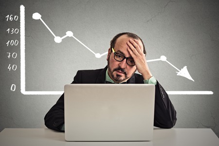 Frustrated stressed business man sitting at table in front of computer with financial market chart graphic going down on grey office wall background. Poor economy concept. Face expression, emotionの写真素材