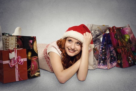 Happy woman in red santa claus hat with christmas gifts dreaming fun holidays. Young girl looking up smiling with shopping bags isolated grey wall background with copy space. Positive face expressionの写真素材