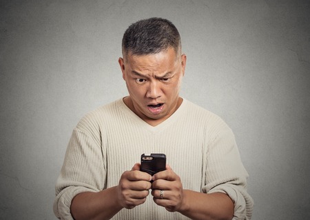 Closeup portrait middle aged surprised stunned shocked pissed off man unhappy by what he sees on cellular phone isolated grey wall background. Negative human emotion face expression feeling reactionの写真素材
