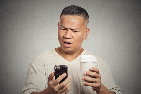 Closeup portrait serious worried middle aged man reading bad news on smart phone holding mobile drinking cup of coffee isolated grey background. Human face expression emotion. Complicated technologyの写真素材
