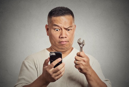 Business idea solution tools concept. Businessman looking at smartphone holding wrench key instrument in hand isolated on office grey wall background. Face expression emotion. New gadget technologyの写真素材