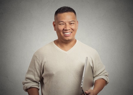 Portrait young handsome happy man holding laptop smiling standing isolated on office grey wall...