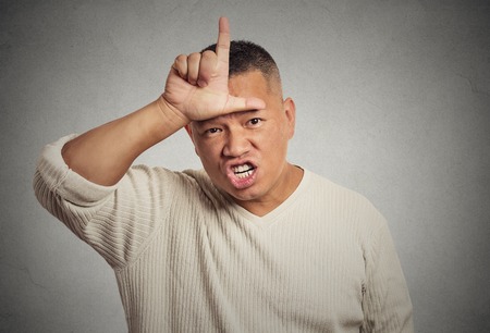 Closeup portrait headshot angry young man showing loser sign on forehead, looking at you with disgust at camera gesture, isolated grey wall background. Negative human emotion facial expression feelingの写真素材