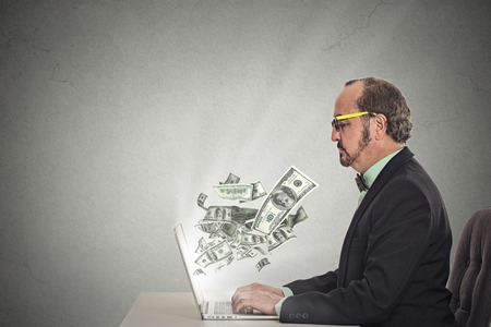 Side profile corporate business man with glasses working online on computer earning money dollar bills banknotes flying out of laptop screen isolated grey wall office background. Human face expressionの写真素材