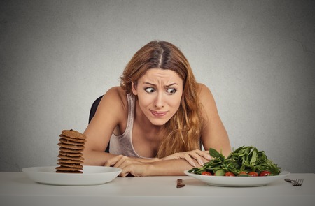 Portrait young woman deciding whether to eat healthy food or sweet cookies she is craving sitting at table isolated grey wall background. Human face expression emotion reaction Diet nutrition conceptの写真素材