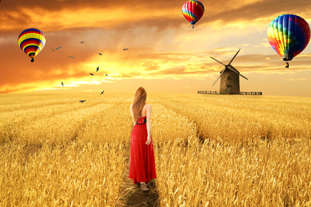Young barefoot woman in red dress standing walking through open wheat field sunrise sunset sky air balloons background. Dreamland nature landscape screen saver. Liberty peace of mind wellness conceptの写真素材