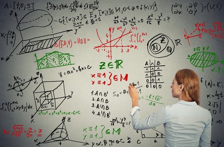 Young businesswoman writing on grey wall complicated formulas text graphic calculations shot from the back. Science education project solution management conceptの写真素材