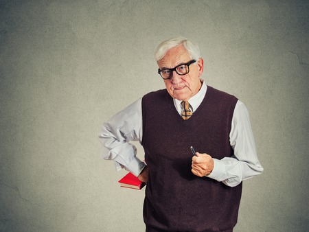 Closeup portrait senior strict teacher holding book and pen, looking very serious, unhappy and grumpy, isolated gray wall background. Human emotion facial expressions. Education conceptの写真素材