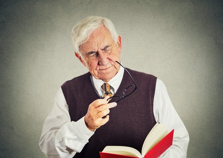 Closeup portrait senior elderly man holding book, glasses having eyesight problems unable to read isolated gray wall background. Human emotion facial expressions. Age related changesの写真素材