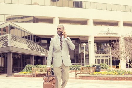 Portrait of young busy happy, smiling business man talking on his phone while walking outside down the street holding briefcase isolated on background of a city. Corporate successの写真素材