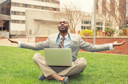 Young businessman with computer relaxing outside corporate office sitting on green grass in...