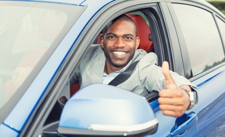 Car. Man driver happy smiling showing thumbs up coming out of blue car side window on outside parking lot background. Young man happy with his new vehicle. Positive face expressionの写真素材