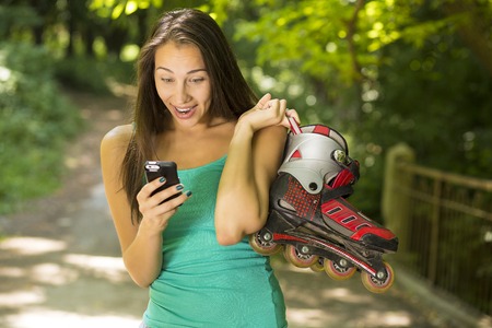 Closeup portrait young surprised woman with inline skates reading news on mobile phone outside on...