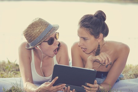 Closeup portrait two surprised girls looking at pad discussing latest gossip news. Young shocked funny women friends reading sharing social media news on mobile pad computer outdoors in parkの写真素材