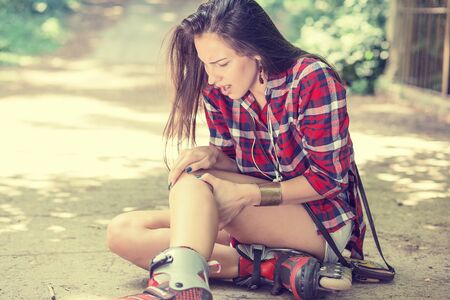 in-line skating injured young woman suffering from pain sitting on the ground touching painful knee...