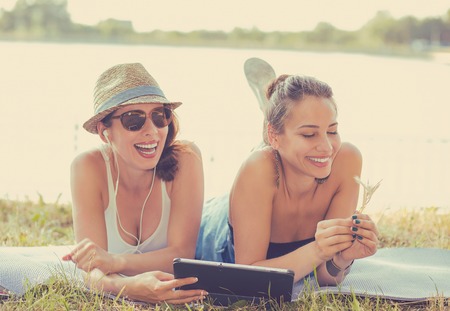 Two funny happy young women friends laughing browsing watching social media videos blogs listen to music on smart pad computer laying outdoors on green meadow on sunny summer dayの写真素材