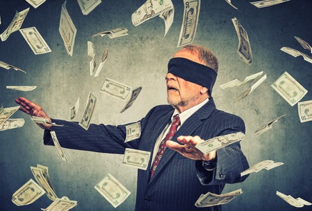 Blindfolded senior businessman trying to catch dollar bills banknotes flying in the air on gray wall background. Financial corporate success or crisis challenge conceptの写真素材