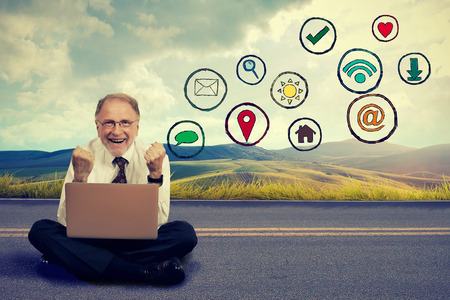 Happy elderly man traveling working on computer using social media application. Smiling executive sitting with laptop on countryside roadの写真素材