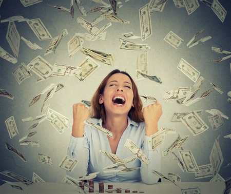 Excited happy young woman sitting at table with growing stack of coins under a money rain isolated on gray wall background. Positive emotions financial success luck good economy concept