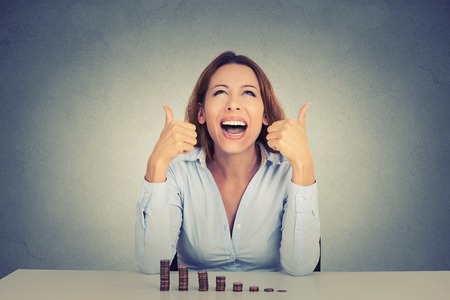 Excited happy young woman sitting at table with growing stack of coins showing thumbs up isolated on gray wall background. Positive emotions financial success luck good economy conceptの写真素材