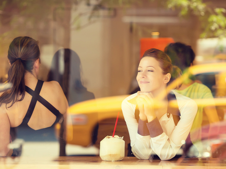 Cafe city lifestyle woman drinking coconut juice, looking outside on street through glass window, sitting indoor juice bar. Young modern female model in her 20s. Positive face expression, emotionの写真素材