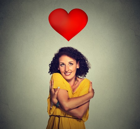 Closeup portrait smiling woman holding hugging herself with red heart above head isolated gray wall background. Positive human emotion, facial expression feeling, attitude. Love yourself conceptの写真素材