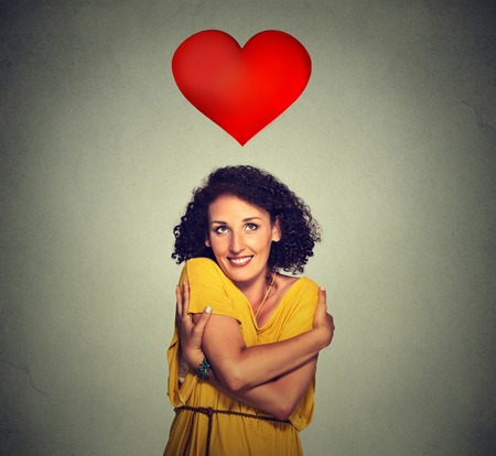 Closeup portrait smiling woman holding hugging herself with red heart above head isolated gray wall background. Positive human emotion, facial expression feeling, attitude. Love yourself concept